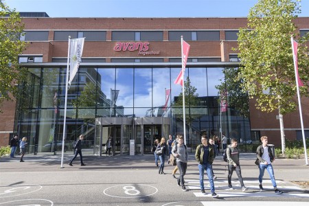 Voorkant van een gebouw van Avans Hogeschool met glazen entree en bakstenen gevel. Op de voorgrond lopen meerdere studenten over het terrein, met vlaggen en bomen langs de ingang.
